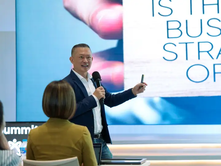 A speaker presenting on stage with slides behind